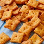 A pile of orange, baked homemade Cheez-Its, made with real extra-sharp cheddar cheese, sit on a blue table.