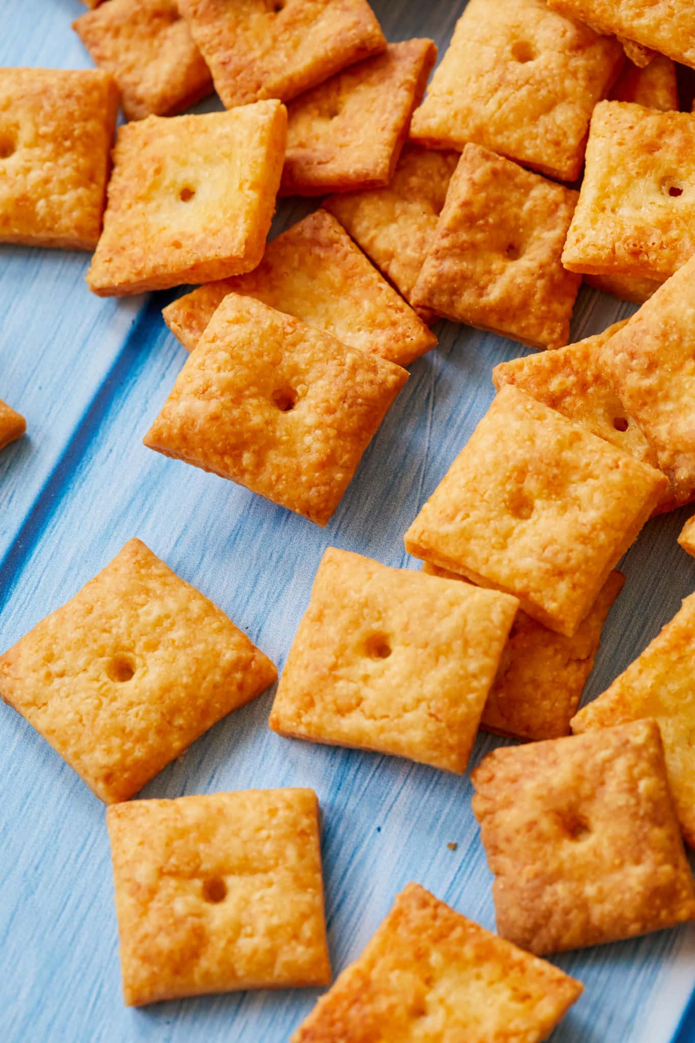 A close-up image of homemade Cheez-Its made with real cheese. They are golden orange with one hole in the center. They are on top of a blue table. 