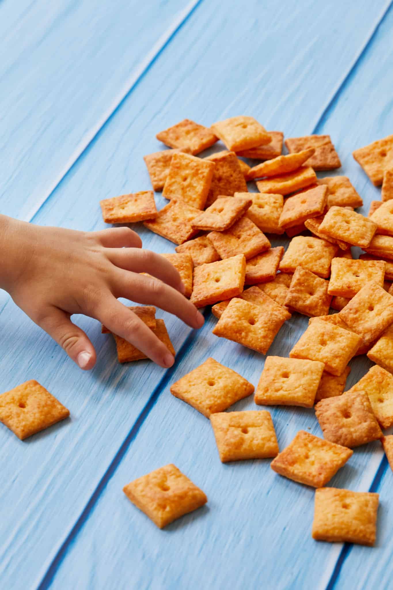 A child's hand reaches out to grab a few homemade Cheez-Its from a pile. The cheesy crackers are orange with a hole in the center.