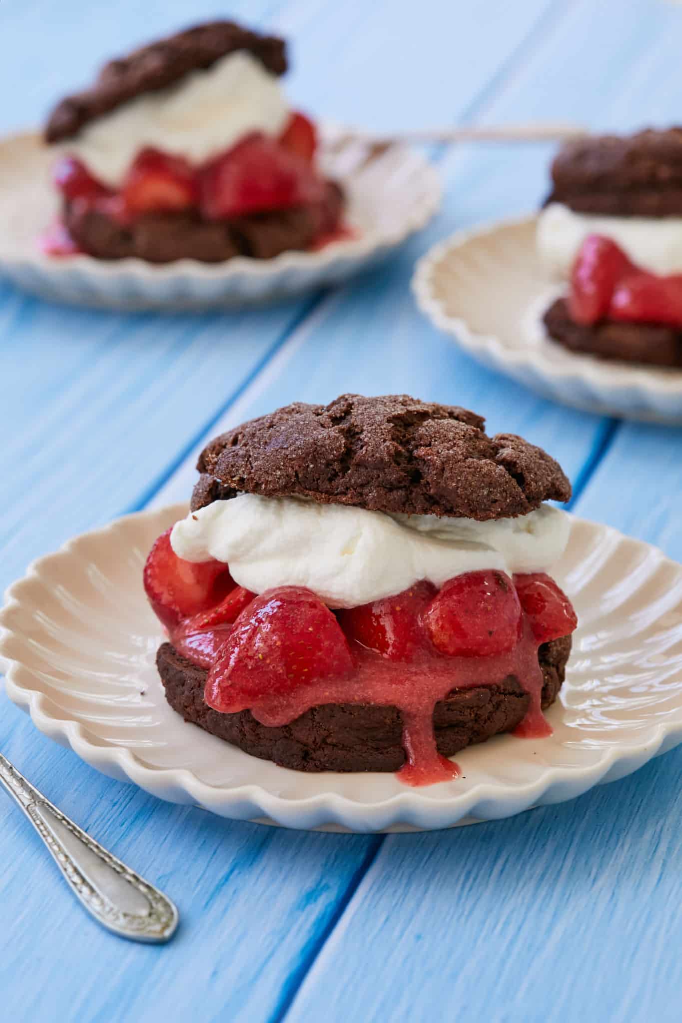 Three chocolate shortcakes filled with whipped cream and macerated strawberries on white plates set on a blue table. 