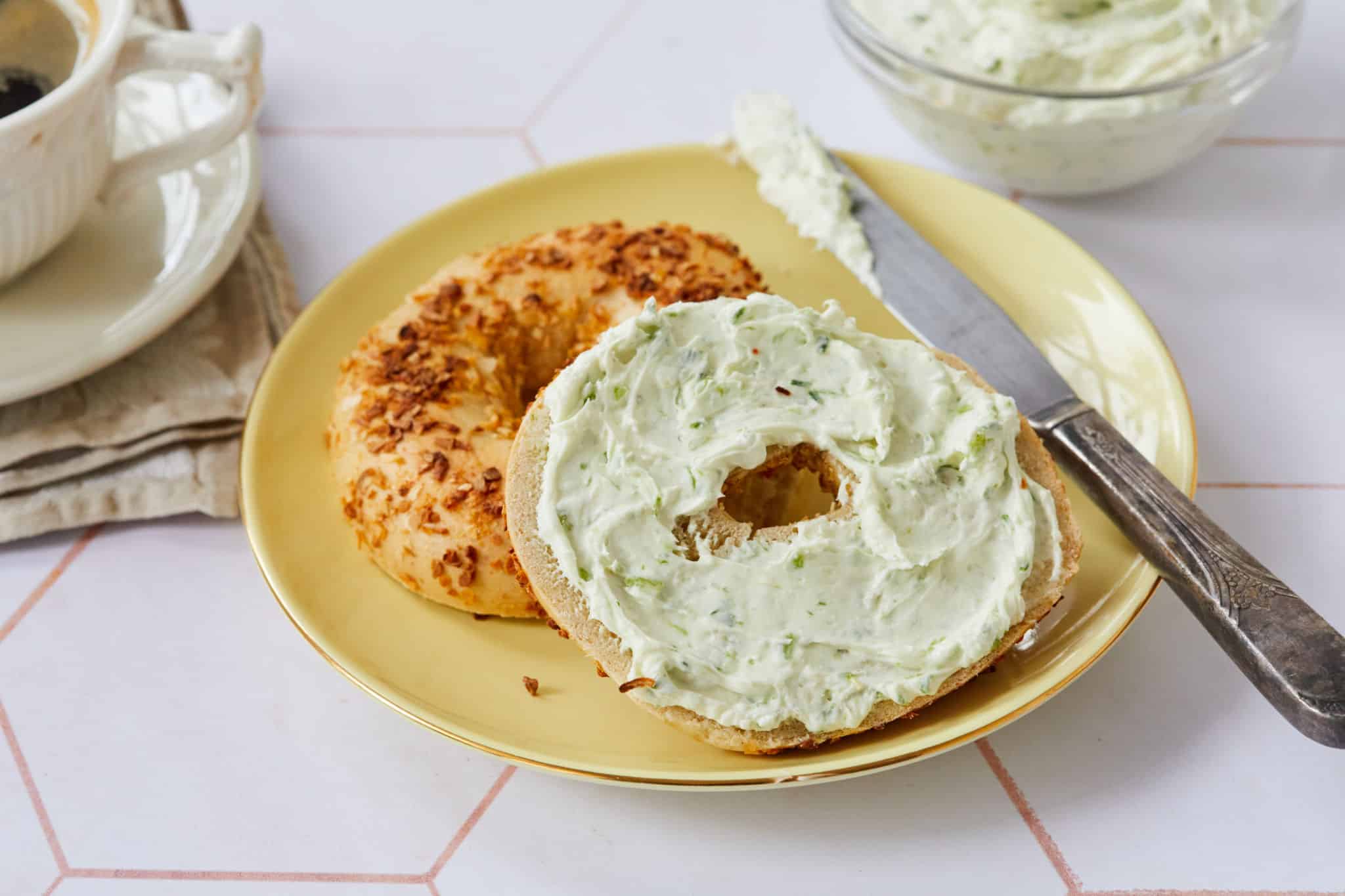Fresh, homemade garlic and chive cream cheese is spread on an everything bagel, served on a yellow plate next to a cup of coffee.