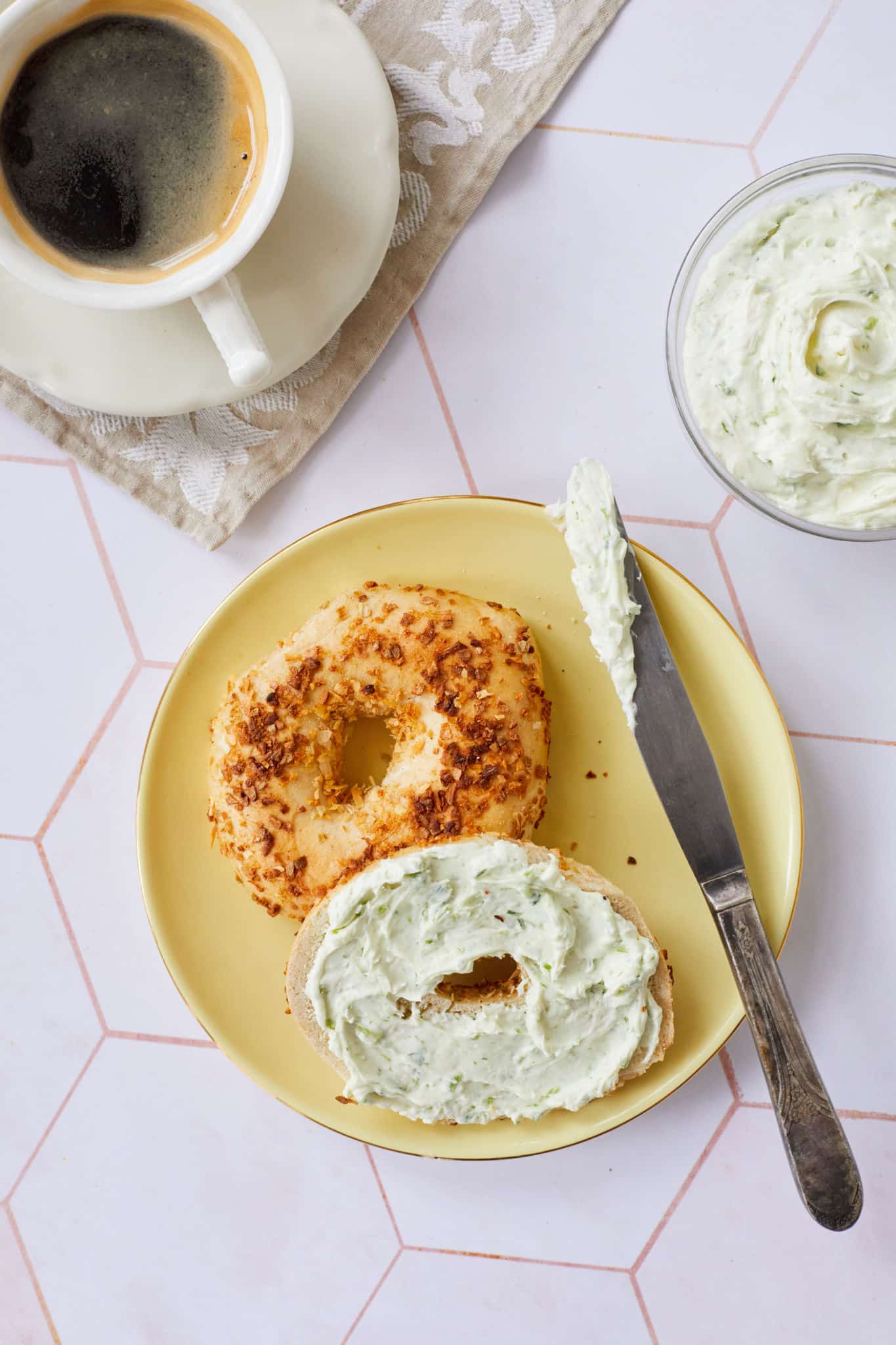Garlic and Chive Flavored Cream Cheese is spread on a garlic bagel, served on a yellow plate, along with a cup of espresso coffee.