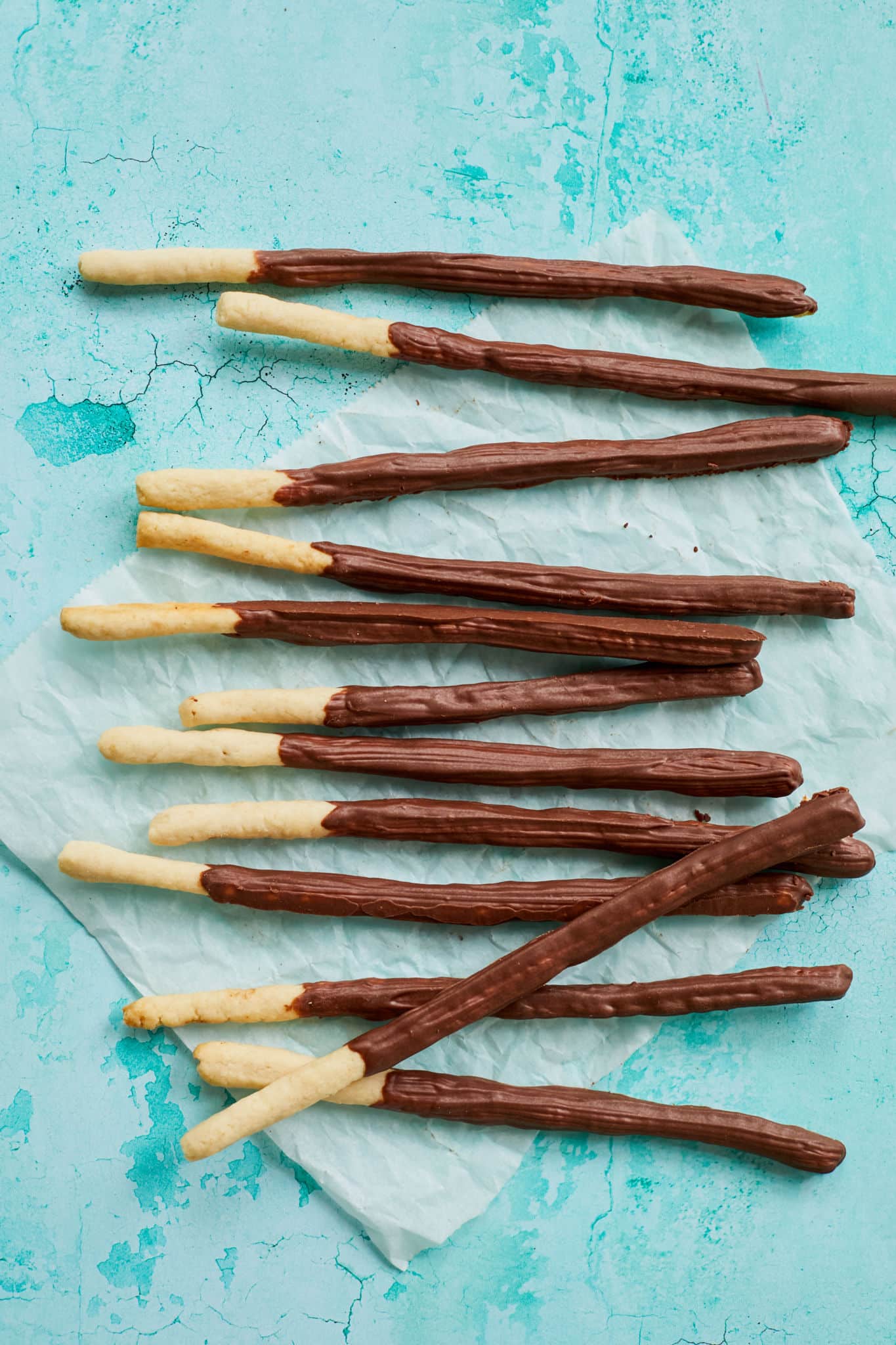 Homemade Pocky sticks are in a thin elongated-stick shape, with most of the stick covered in a chocolate-flavored coating while leaving a small section uncoated for easy handling. The cookies are placed on parchment paper with a light teal background. 