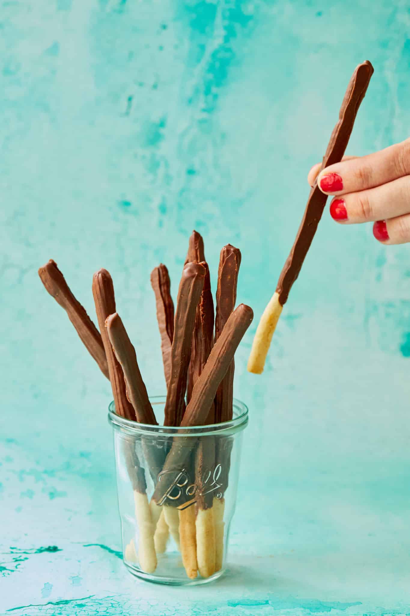 Homemade Pocky sticks are in a thin elongated-stick shape, with most of the stick covered in a chocolate-flavored coating while leaving a small section uncoated for easy handling. The cookies are placed in a glass Ball mason jar with a light teal background. One Pocky biscuit is being taken out of the jar. 