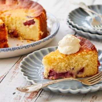 A slice of Italian Ricotta Cake With Plums is served on a blue plate next to the entire cake it has been cut out of, with a dollop of whipped cream.