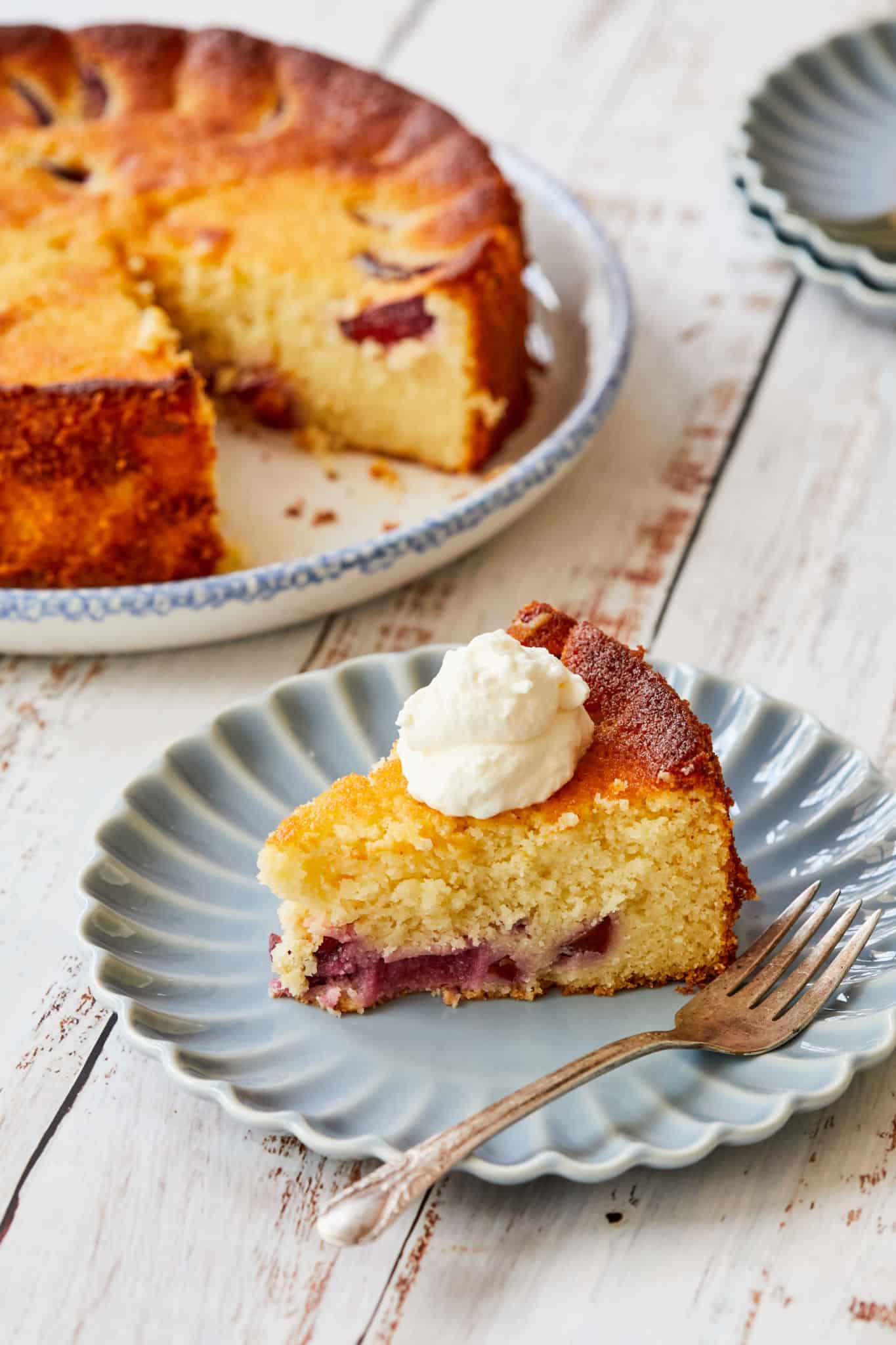 A close-up image of Italian Ricotta Cake which shows the baked black plums. It is served with fresh whipped cream. In the background of the photo is the rest of the circular cake.