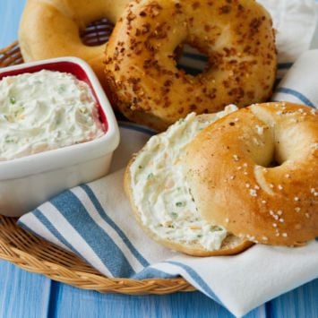 A bagel is topped with homemade jalapeno and cheddar cream cheese and sits in a basket next to two other whole bagels, one plain, one everything bagel, and a dish filled with the homemade cream cheese.