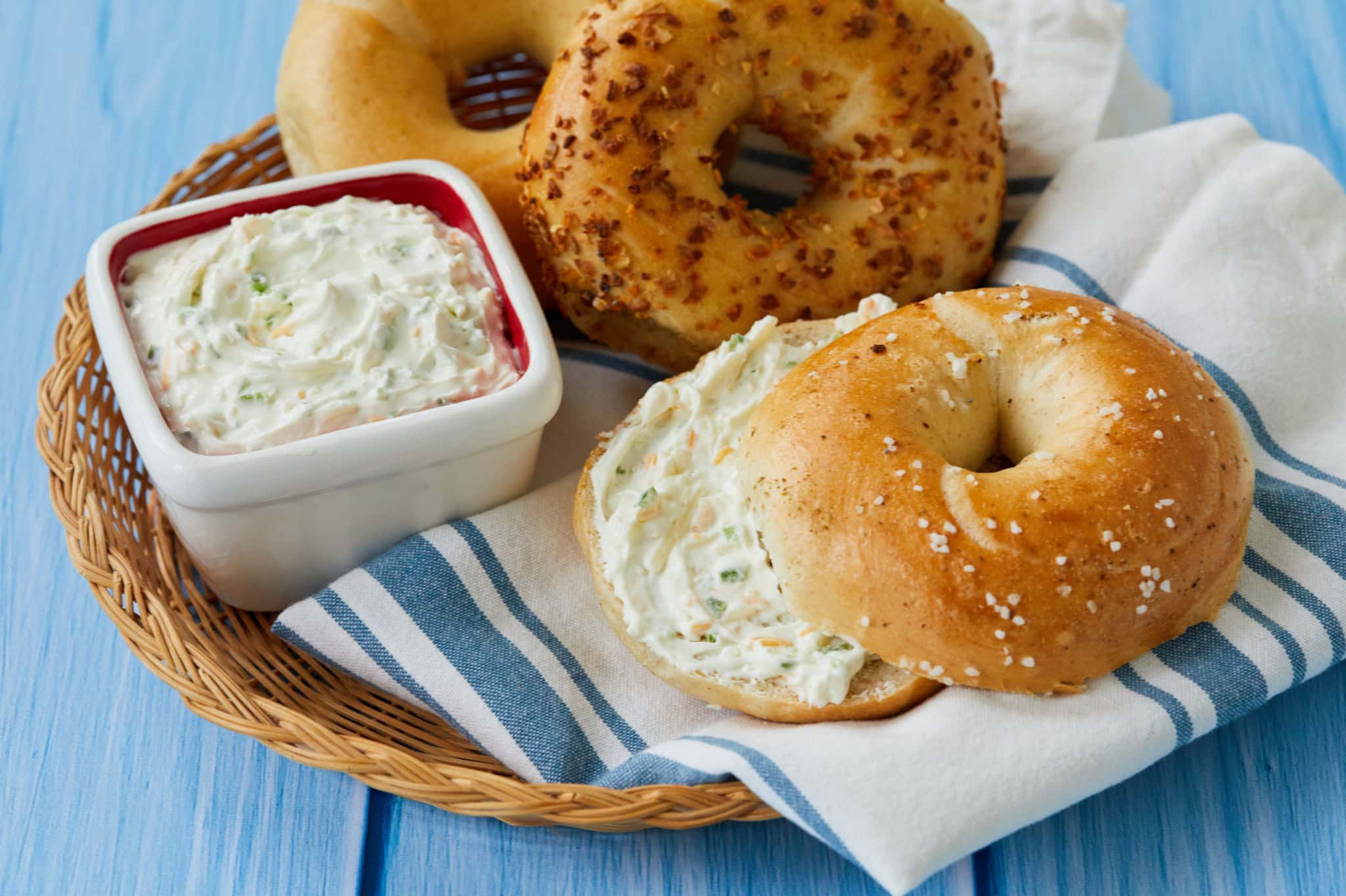 A bagel is topped with homemade jalapeno and cheddar cream cheese and sits in a basket next to two other whole bagels, one plain, one everything bagel, and a dish filled with the homemade cream cheese.