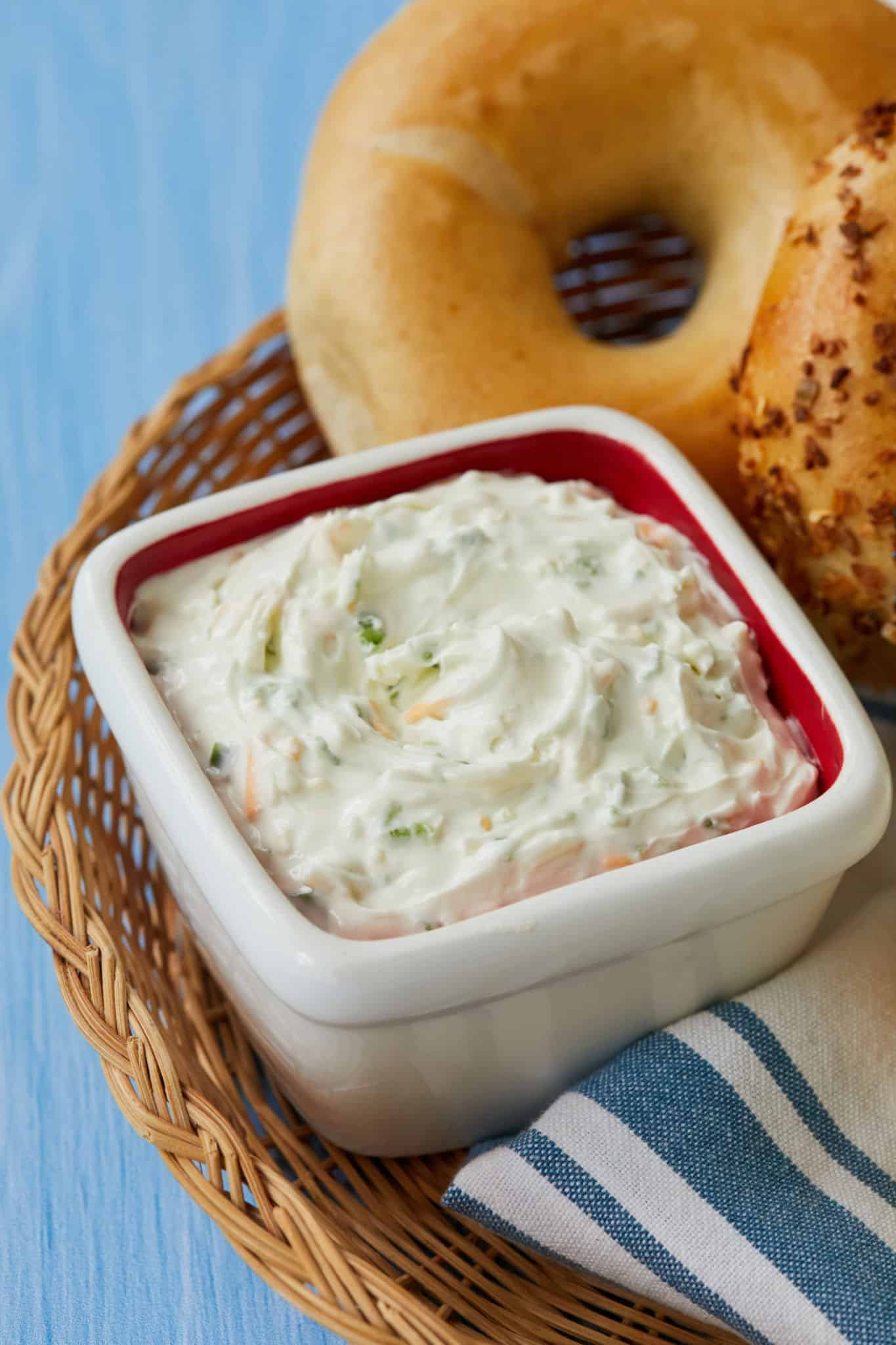 A square white bowl is filled with homemade jalapeno cheddar cream cheese. The cream cheese is whipped with fresh jalapenos. Next to the cream cheese are two bagels: one plain bagle and one everything bagel.