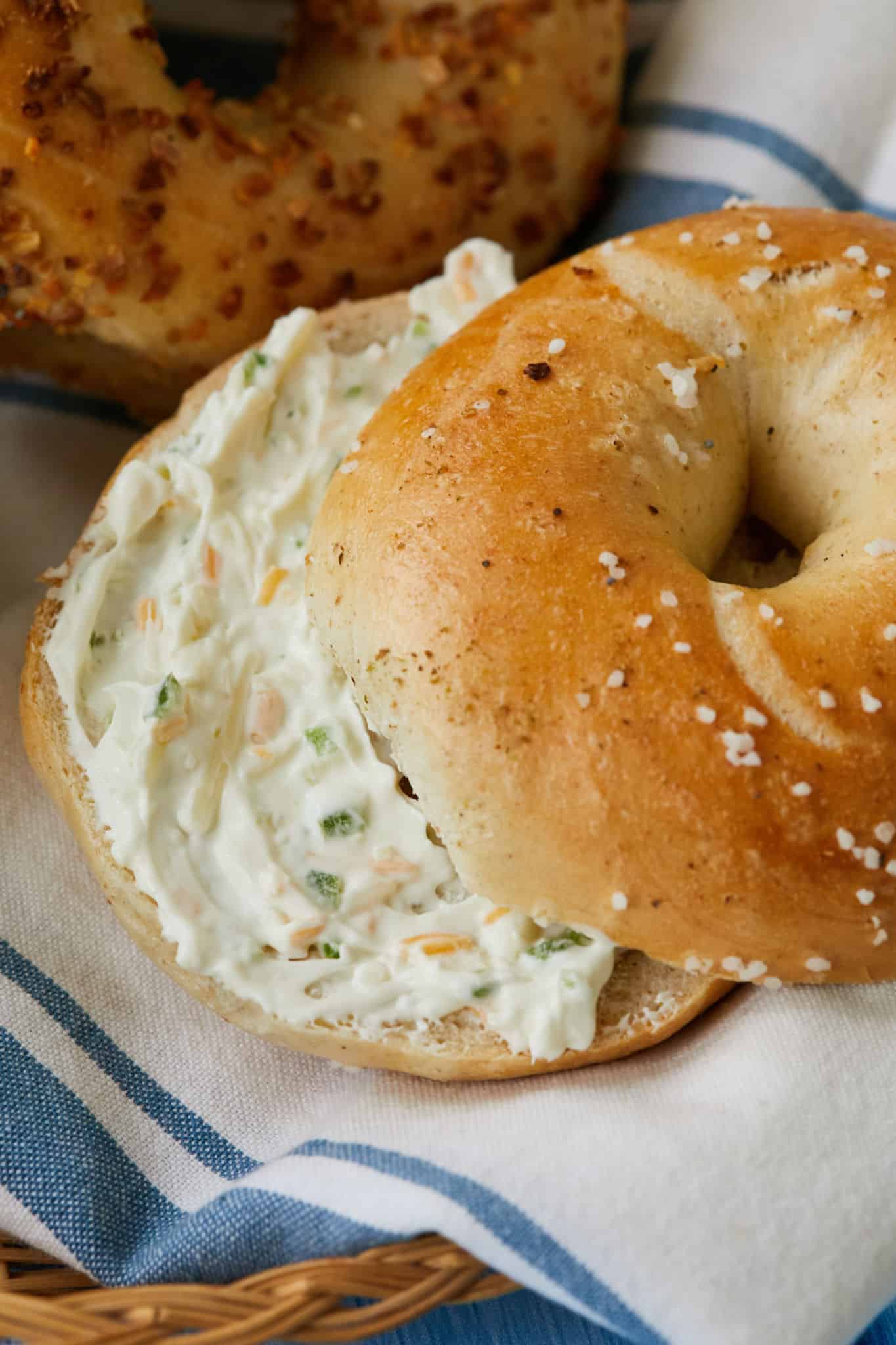 A salt bagel is cut in half and homemade jalapeno cheddar cream cheese is spread in the middle. An everything bagel sits behind the salt bagel.