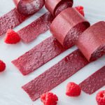 Five individual rolls of raspberry fruit leather are rolled up, exposing just a bit of their end, on a table with raspberries.