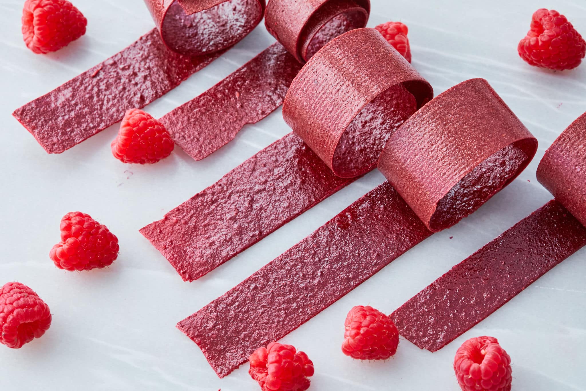 Five individual rolls of raspberry fruit leather are rolled up, exposing just a bit of their end, on a table with raspberries.