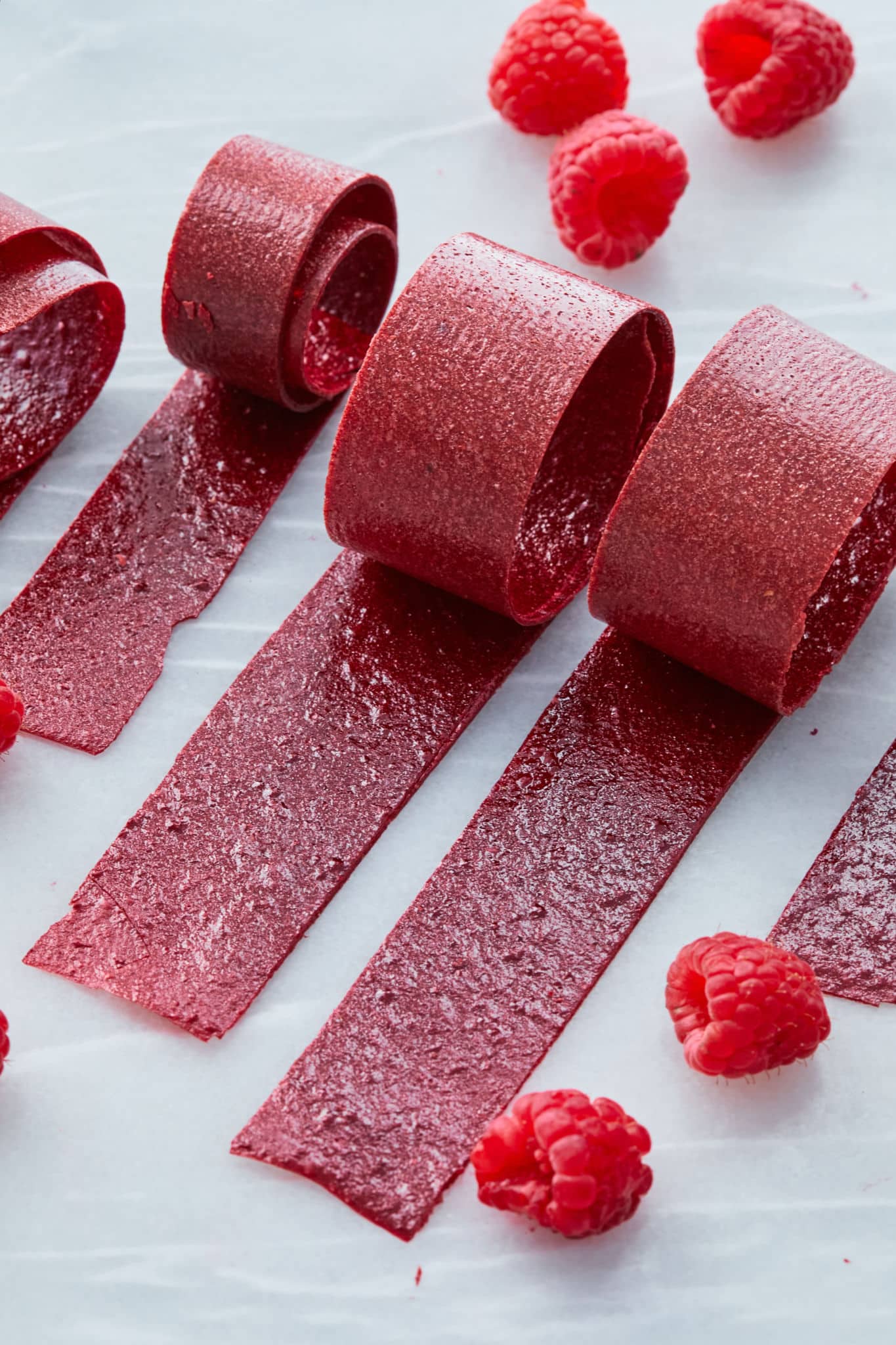 A close up photo of raspberry fruit leather, cut into about an inch-wide strips, are rolled up on a table.