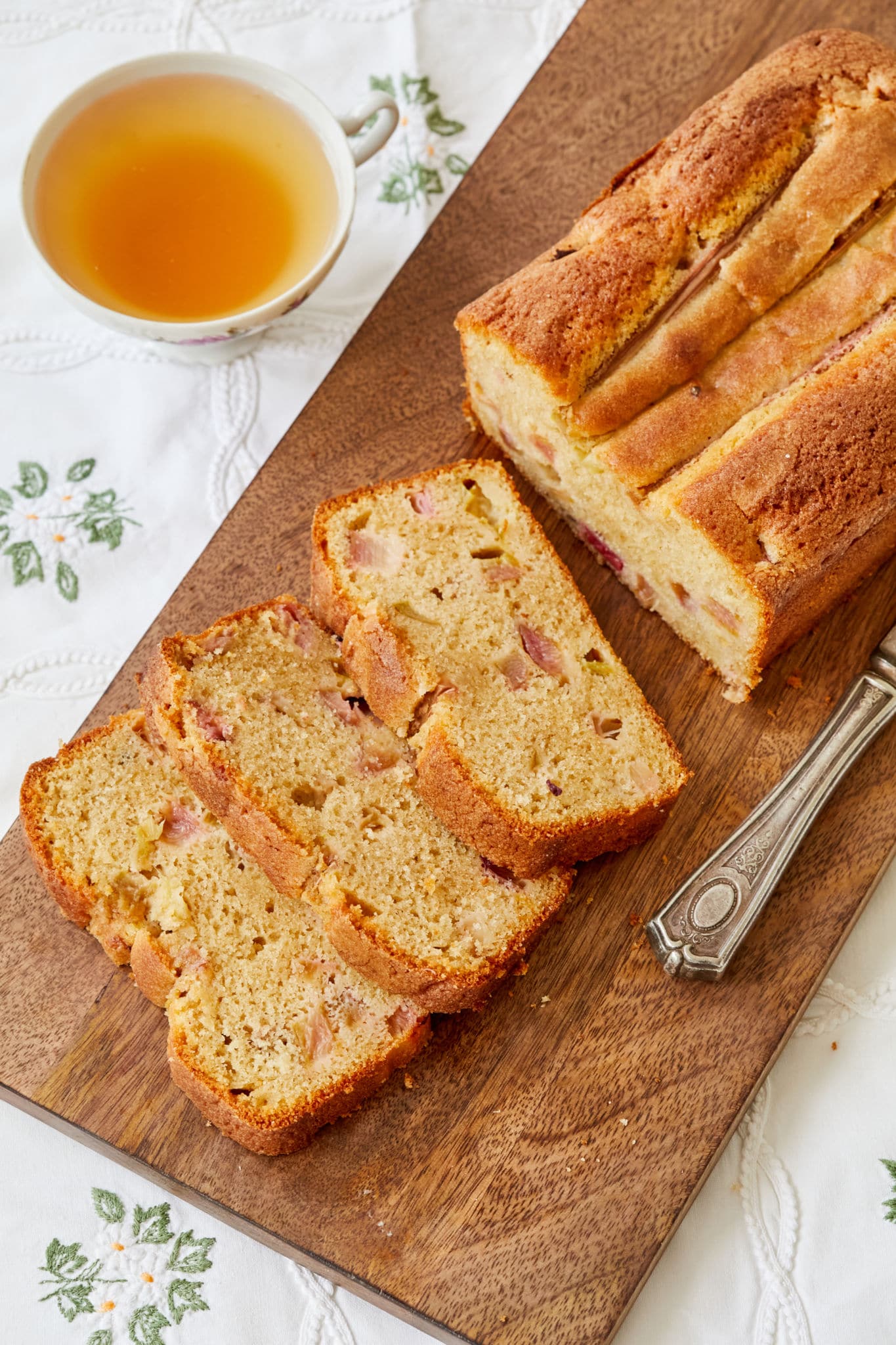 A loaf of rhubarb ginger quick bread is cut into three slices and displayed on a wooden cutting board on top of a white tablecloth and next to a cup of tea.