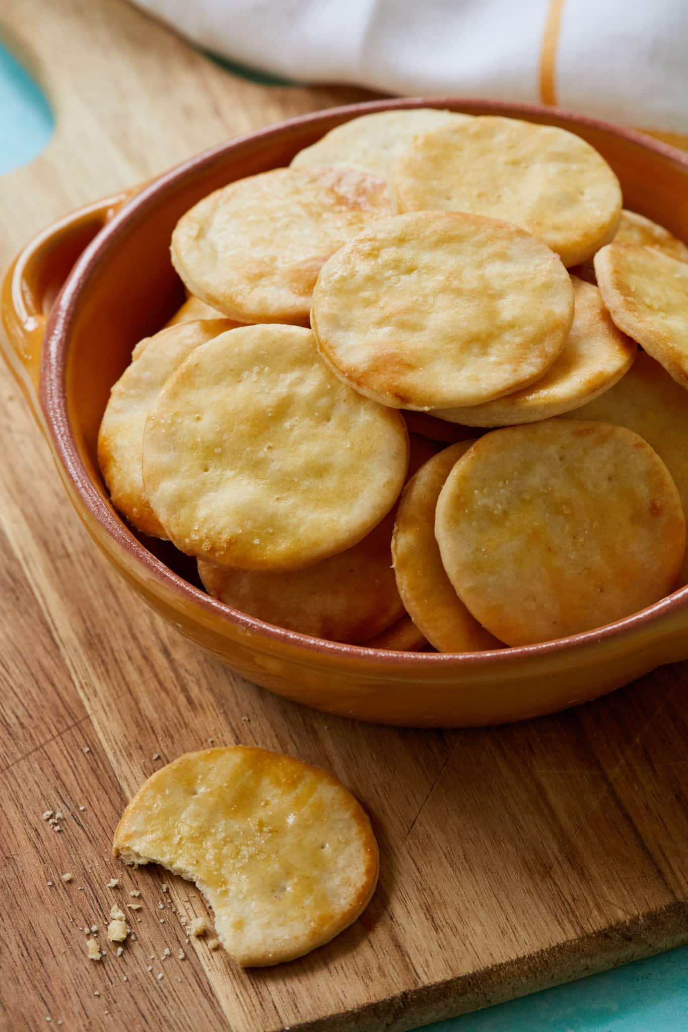 A close up of crispy, buttery homemade Ritz Crackers. The crackers are in a bowl, one is on a wooden cutting board with a bite taken out of it.