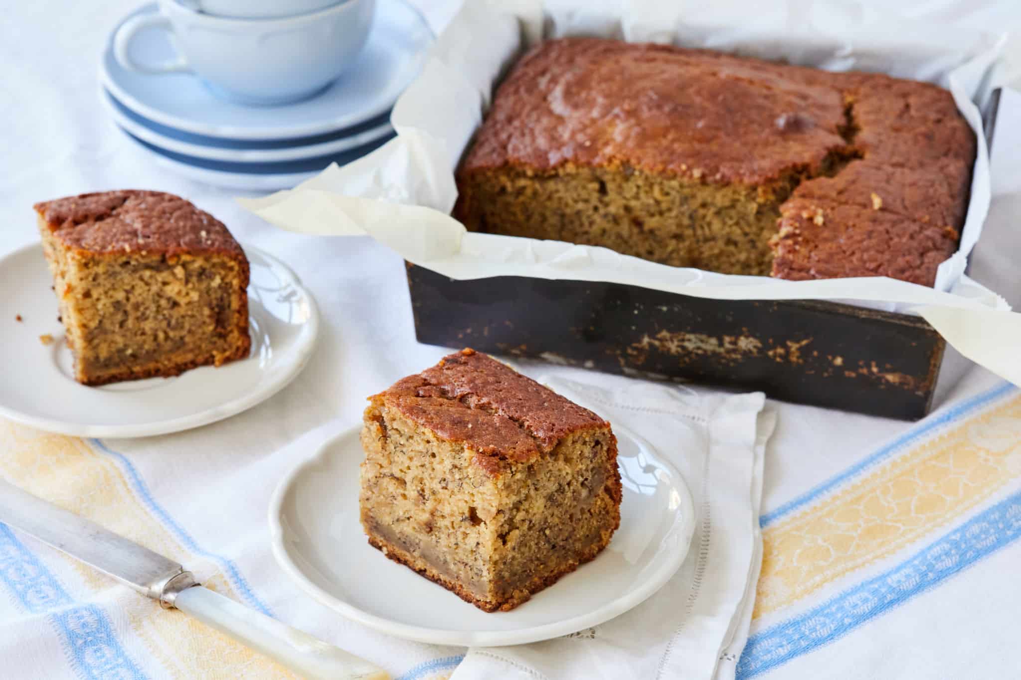 Two square slices of homemade sourdough banana bread are served on plates next to the loaf in the baking pan.