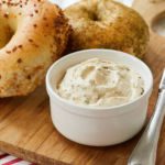 Homemade Sun-Dried Tomato Cream Cheese is served in a white dish next to a garlic bagel and a poppyseed bagel.