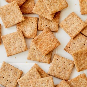 A number of crispy homemade Wheat Thins are spread out on a white background.