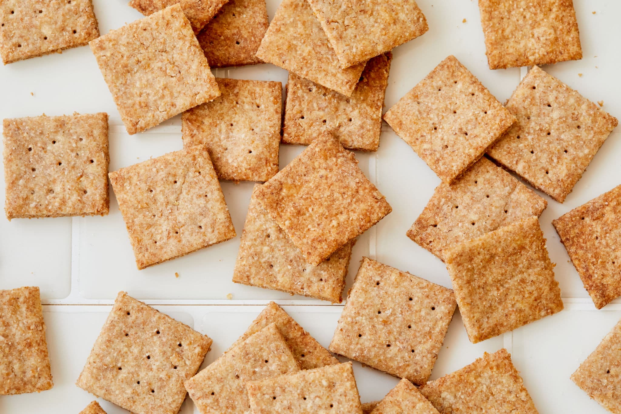 A number of crispy homemade Wheat Thins are spread out on a white background.