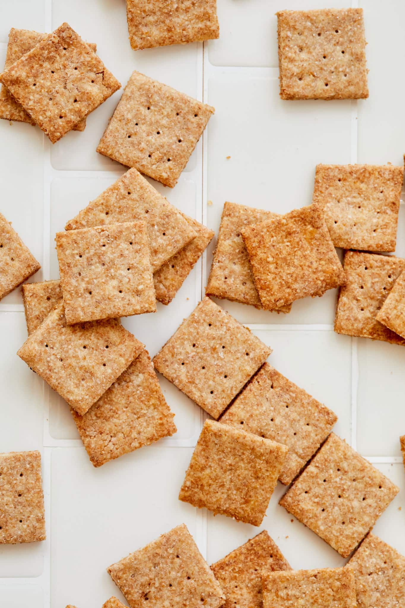 A number of homemade wheat thins, which are evenly sized and baked, are on top of a white gridded background.