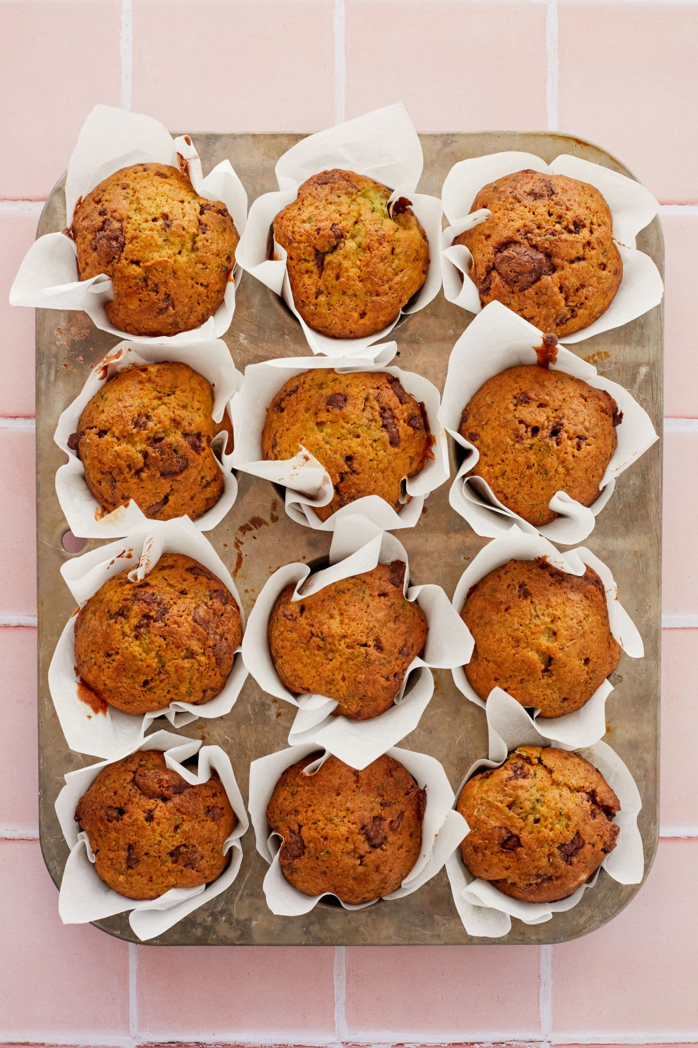 A dozen zucchini muffins with chocolate chips rest in their baking tray on top of a pink tile background.