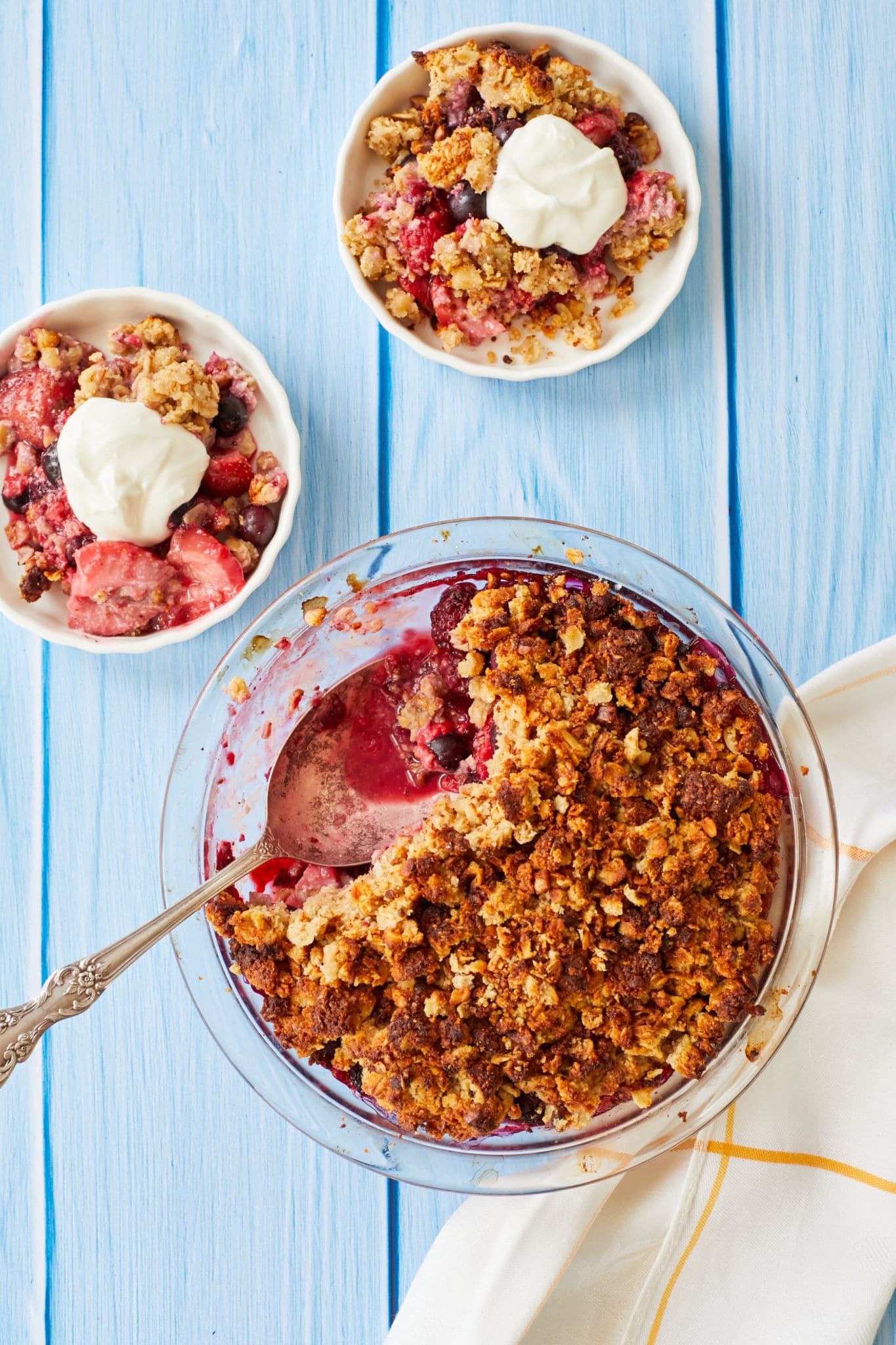 A photo of healthy berry crisp. There are two servings in white bowls next to the main dessert in a clear glass pie tin. Scoops of vanilla Greek yogurt are on top of the crisp.