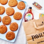 A dozen chewy maple pumpkin cookies on a baking sheet next to a grocery bag full of ingredients.