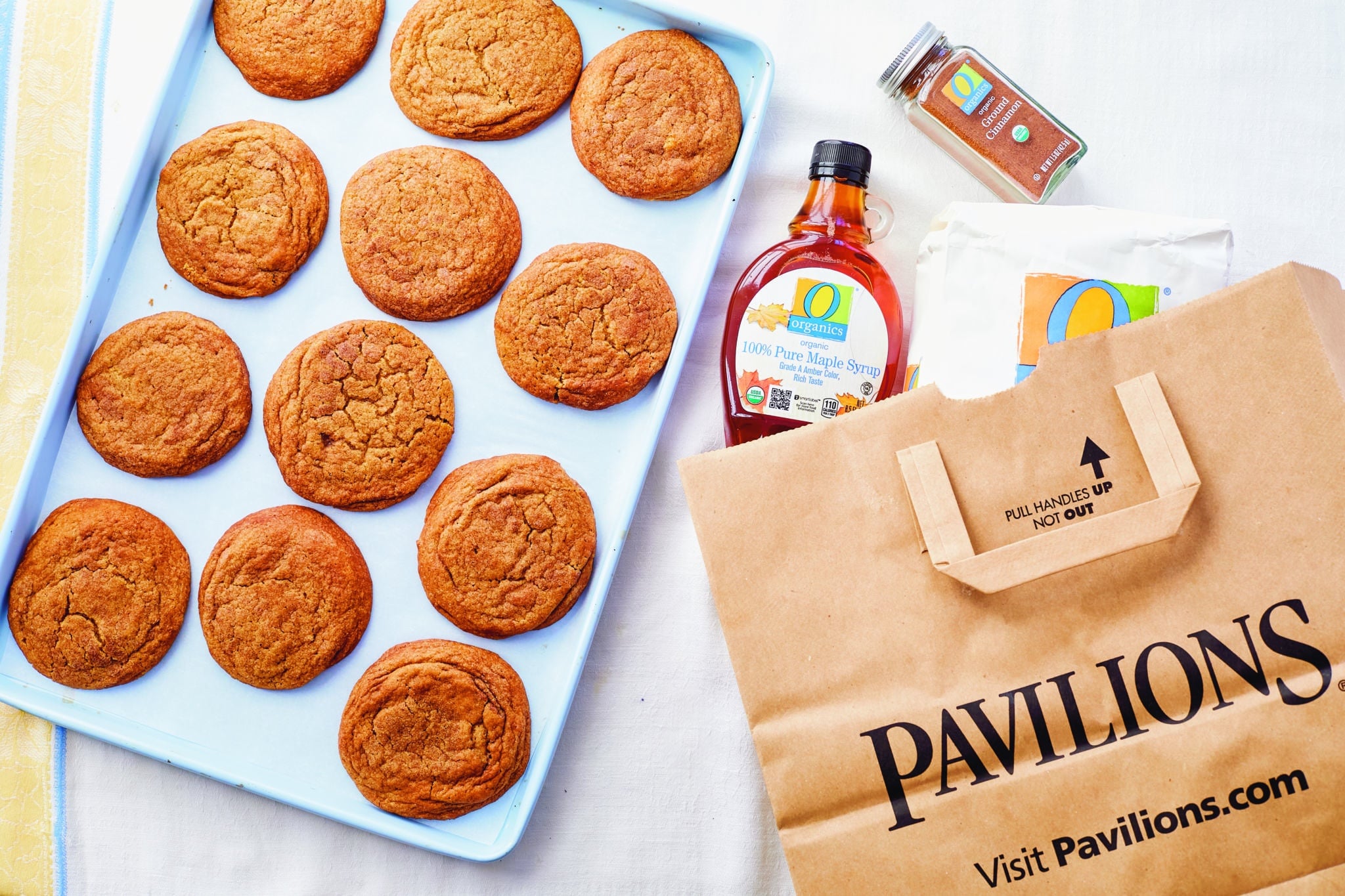 A dozen chewy maple pumpkin cookies on a baking sheet next to a grocery bag full of ingredients.