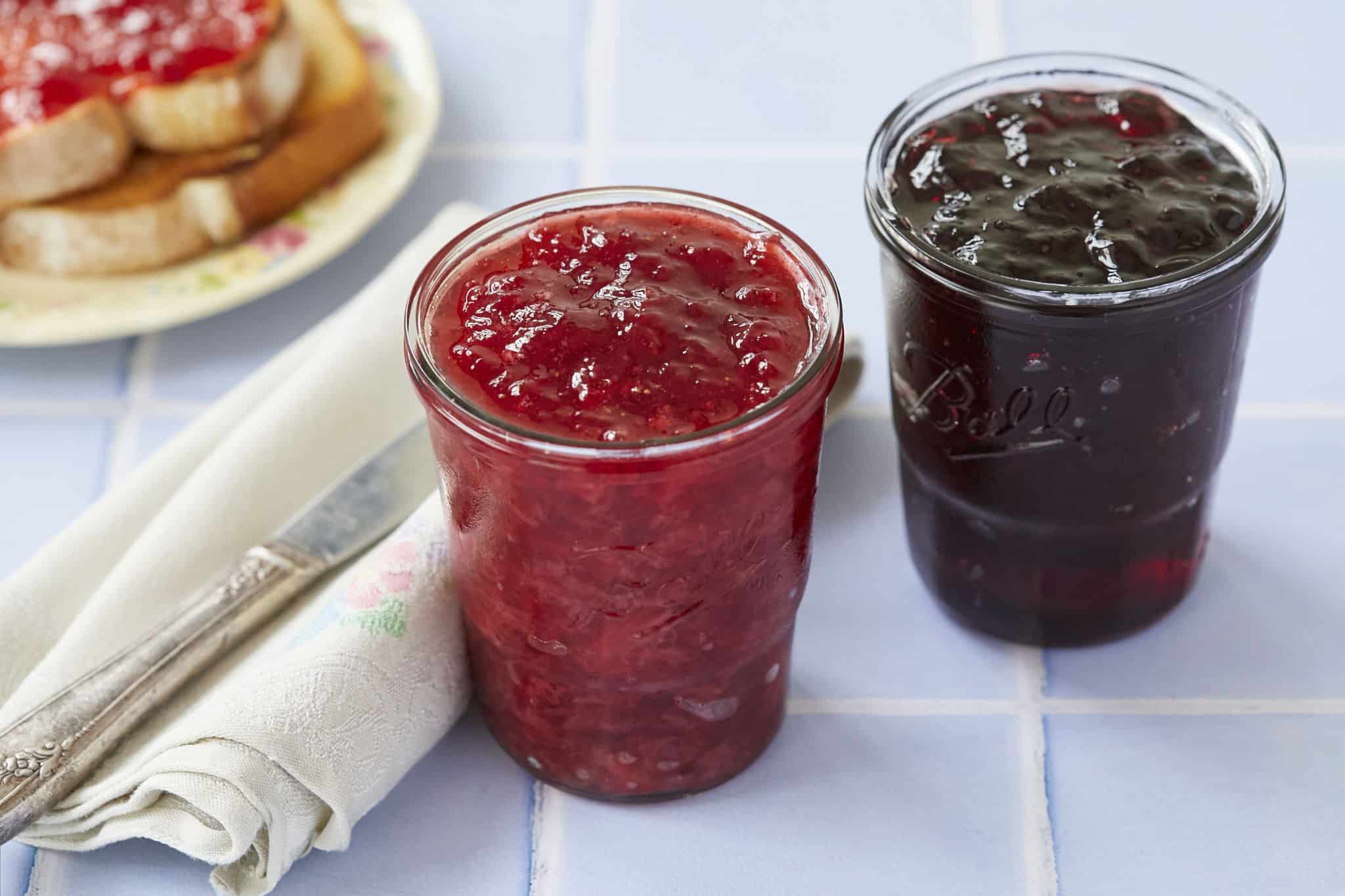 Two homemade jars of jam are displayed on a table, one red and one dark purplish. Possibly homemade raspberry or strawberry jam.