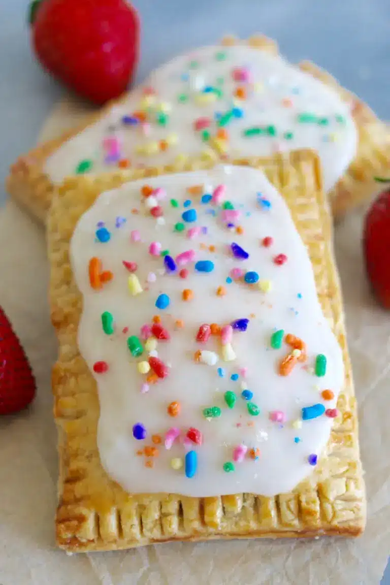 Two homemade Pop-Tarts are on a table. These homemade strawberry Pop-Tarts are next to real strawberries and topped with rainbow sprinkles. 