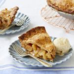 A slice of Apple Cheddar Pie is served on a dish with a scoop of vanilla ice cream. The apple pie filling is topped with a cheddar cheese crust.