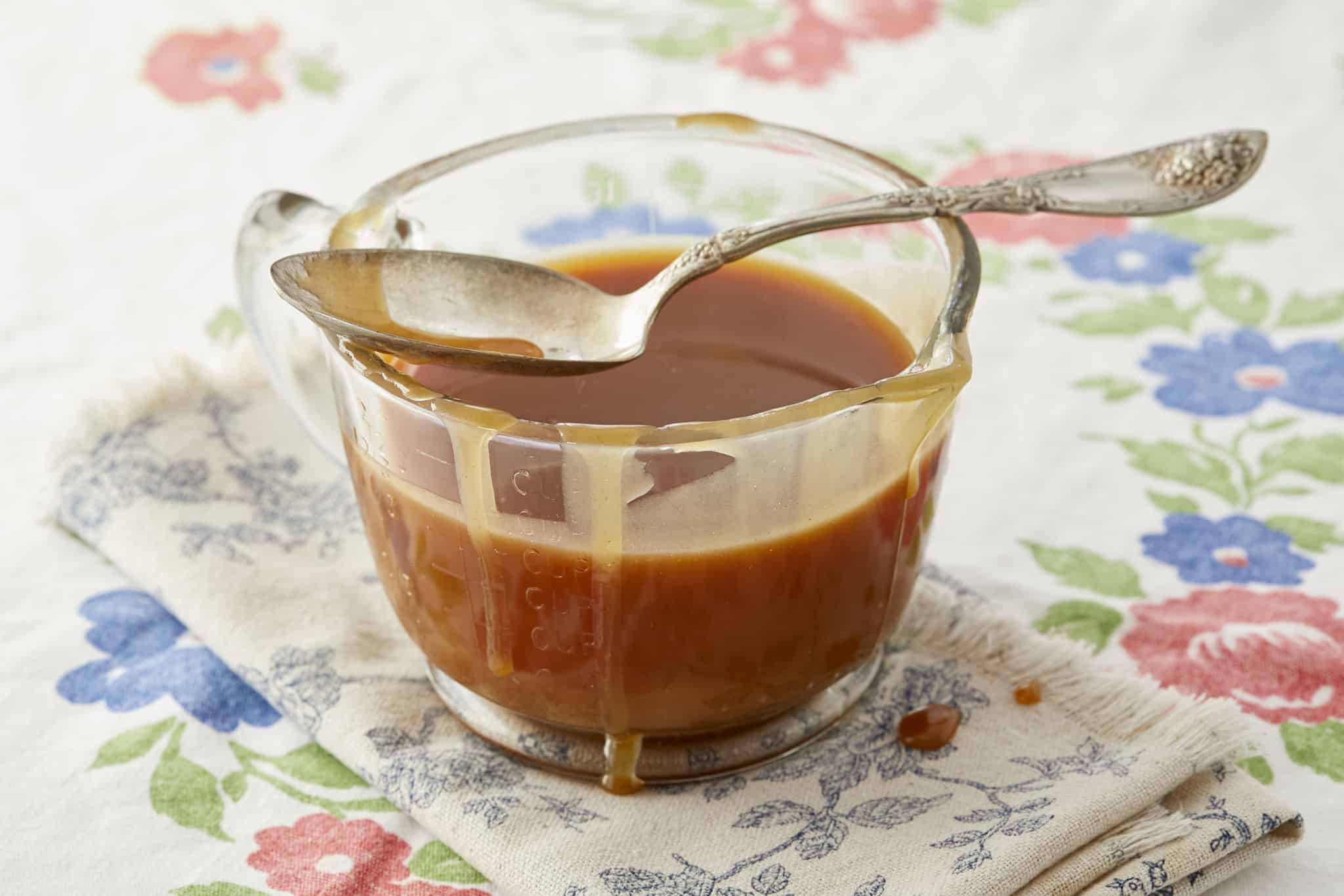A glass jar of apple cider caramel sauce, with a spoon laid across the top, on a floral tablecloth.