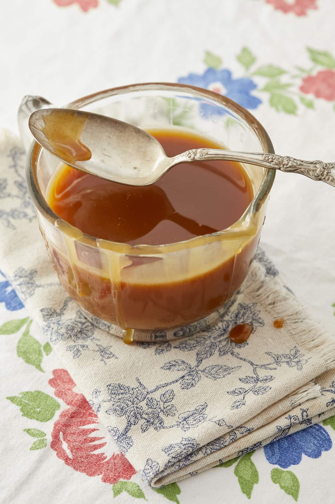 A glass jar full of apple cider caramel sauce, with a spoon laid on the edge, on a linen napkin and tablecloth