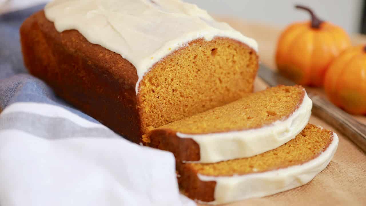 A loaf of pumpkin bread is sliced on a baking tray, next to small pumpkins in the background. It is served with a white glaze.
