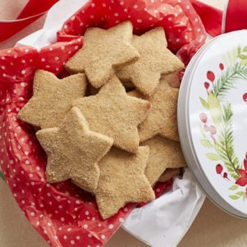 Homemade Biscochitos cut into a star shape and coated in cinnamon sugar are served in a Christmas tin with red tissue paper.