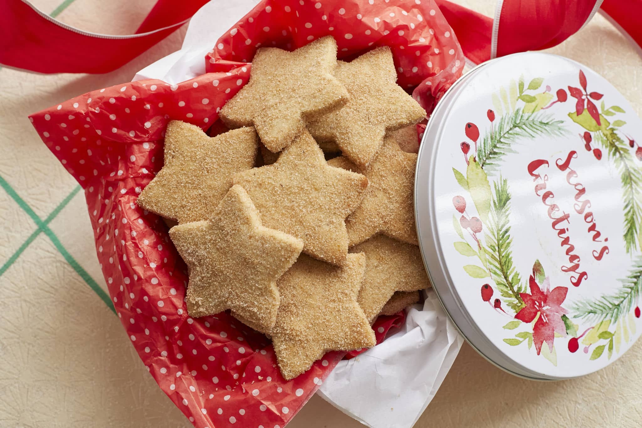 Homemade Biscochitos cut into a star shape and coated in cinnamon sugar are served in a Christmas tin with red tissue paper.