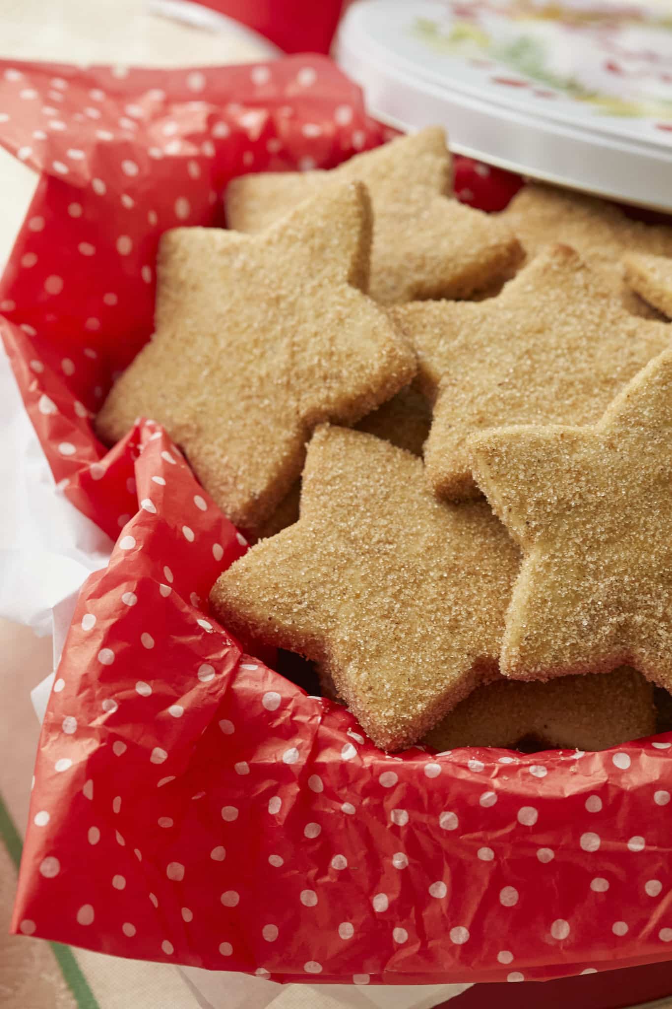 A close-up image of homemade Biscochitos, or traditional New Mexican Christmas Cookies, are presented in a gift tin. The cookies are cut into star shapes and coated in cinnamon sugar.