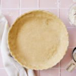 Buttermilk pie crust pressed into a pie tin, next to a glass bowl of flour with a pastry mixer inside and metal measuring cups, laid out on a pink tile counter top.