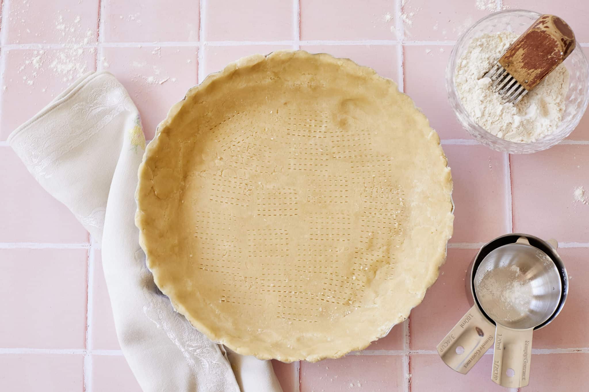 Buttermilk pie crust pressed into a pie tin, next to a glass bowl of flour with a pastry mixer inside and metal measuring cups, laid out on a pink tile counter top.