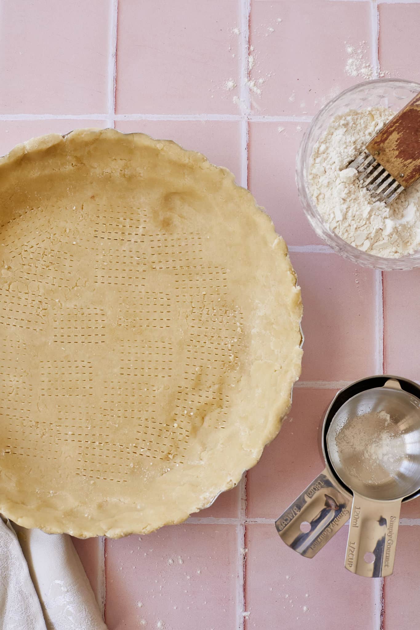 An overhead shot of buttermilk pie crust pressed into a pie tin, on top of a linen napkin, next to a glass bowl of flour and butter with a pastry blender, and metal measuring spoons, on a pink tile counter.