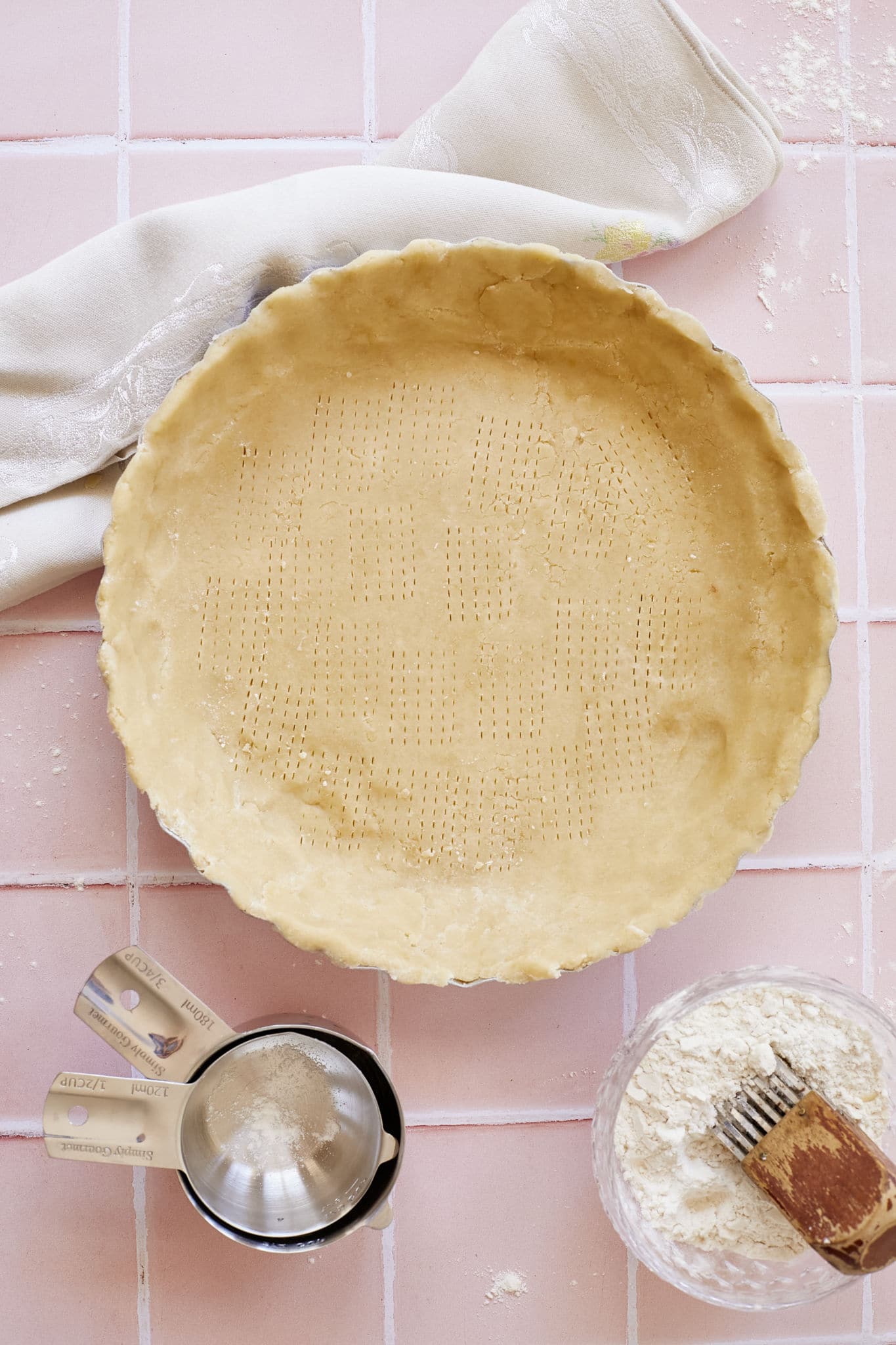 Buttermilk pie crust pressed into a pie tin, with fork marks to let out steam, next to a napkin, glass bowl of flour and butter with a pastry blender, and metal measuring spoons on a pink tile counter.