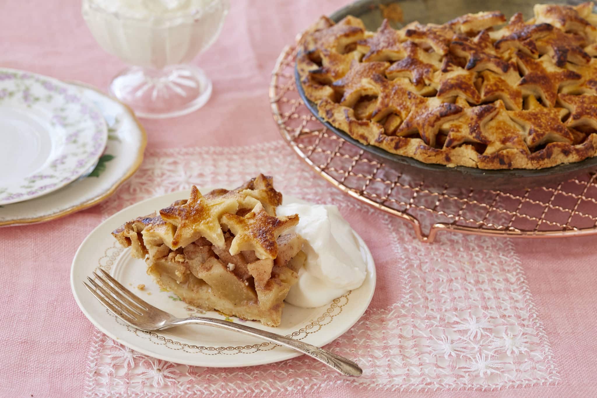 A slice of homemade Pear Buttertscotch Pie is served on a white plate on a pink table cloth. In the upper right corner is the rest of the pear butterscotch pie in the baking tin. To the upper left are two empty plates, stacked, and homemade whipped cream.