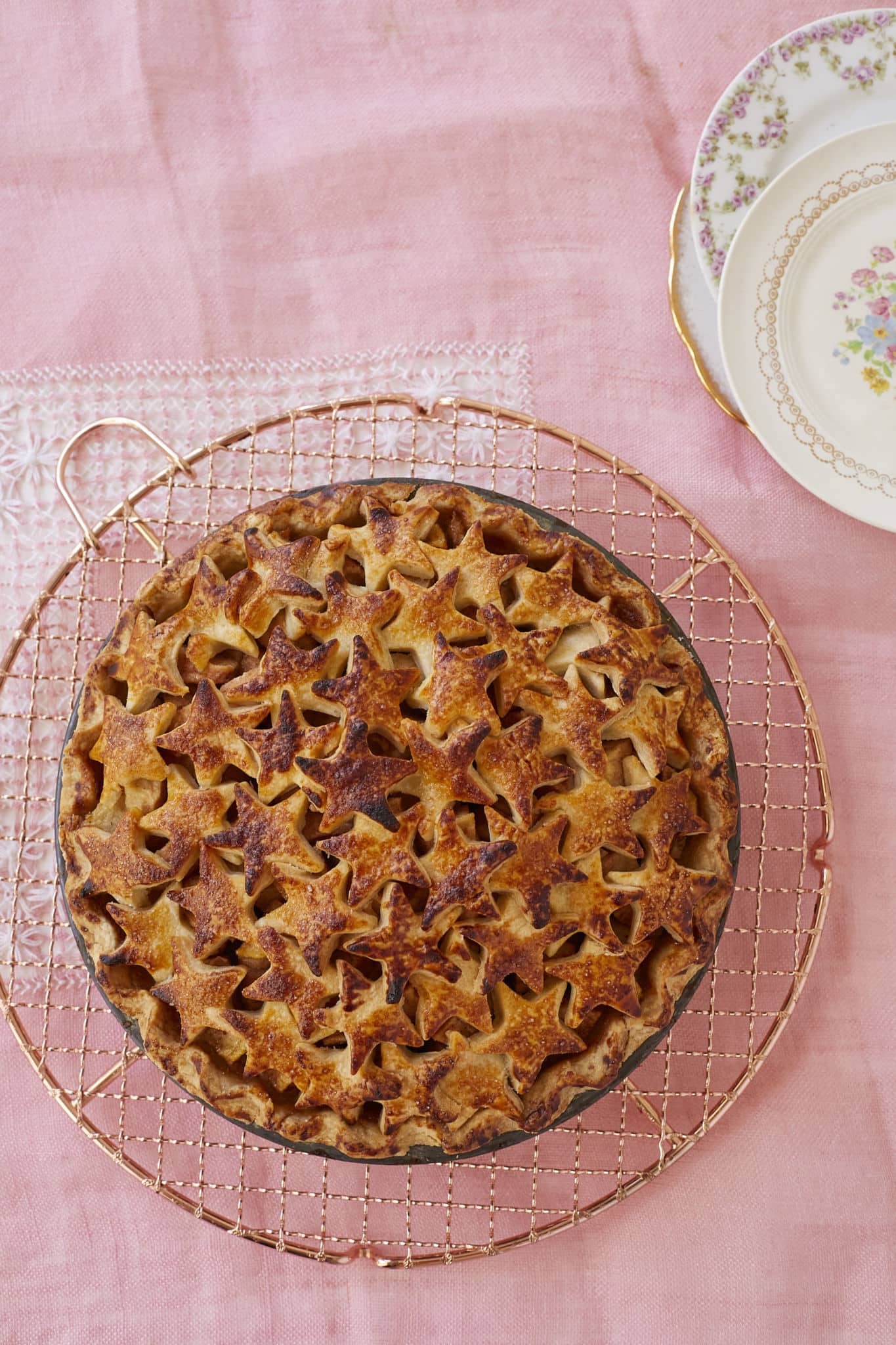 A photo of Butterscotch Pear Pie taken from above. The homemade pear pie is presented on a copper cooling rack on top of a pink table cloth. China dishes are stacked in the top right of the photo.