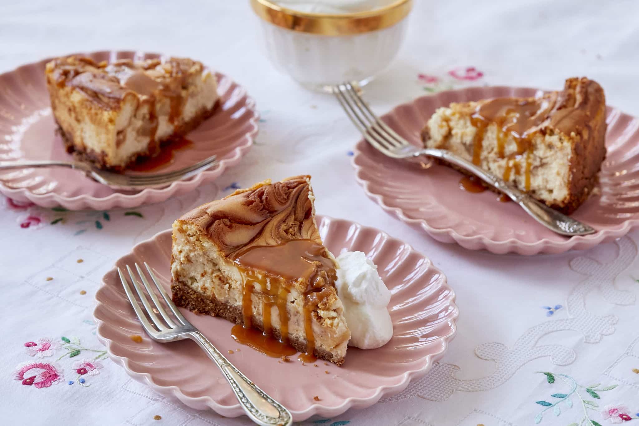 Three slices of homemade Caramel Apple Cheesecake are served on pink plates on a floral table cloth. The cheesecake is moist and jiggly with melted caramel dripping down the sides.
