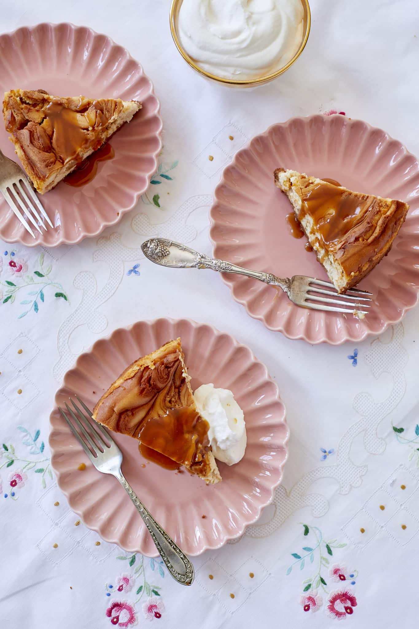 Apple Caramel Cheesecake slices are served on pink plates with homemade salted caramel sauce and whipped cream