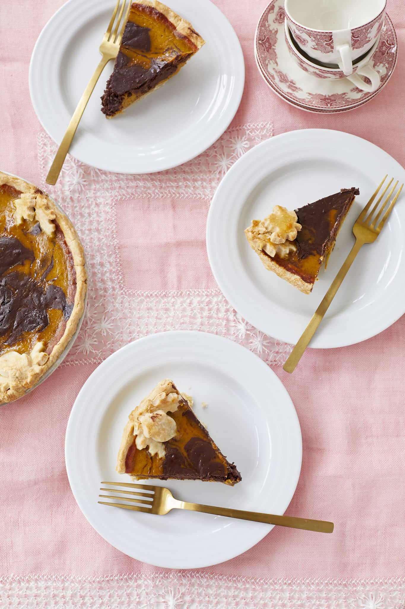 Three slices of Chocolate Swirl Pumpkin Pie are served on white plates with gold forks. The photo, taken from above, shows the decorative pie crust and the marbled pie filling.