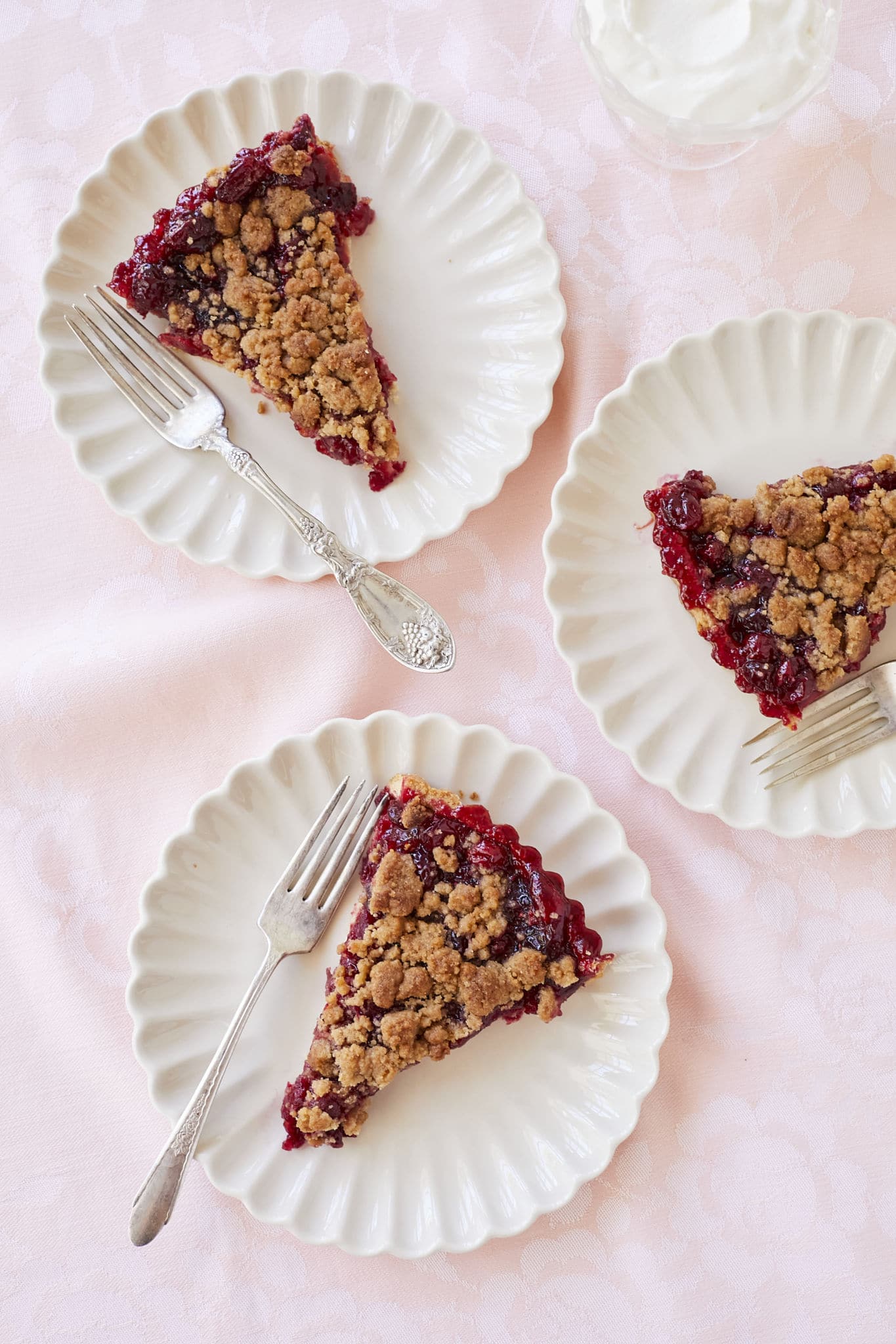 Three slices of Cranberry Tart With Orange are served on white plates. The rich red coloring of the cranberry filling is visible underneath the brown sugar crumble topping. 