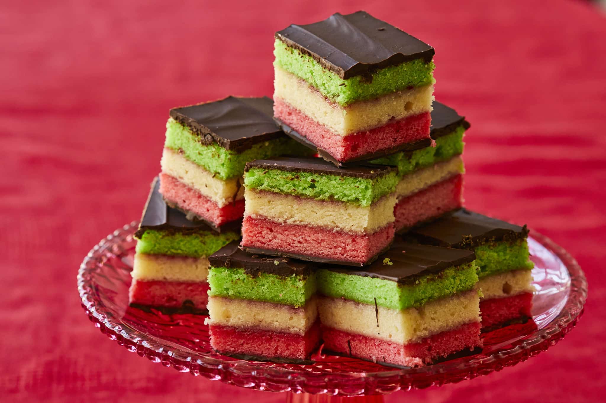 Homemade Italian Rainbow Cookies are stacked on a tray. The tricolor cookies are pink, white, and green, and coated in semi-sweet chocolate.