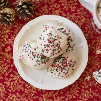 A plate of homemade Italian Ricotta Cookies are presented on a red table cloth with gold accents next to a couple of pine cone. The Italian Ricotta Cookies are topped with a homemade vanilla glaze and red, green, and white Christmas sprinkles.