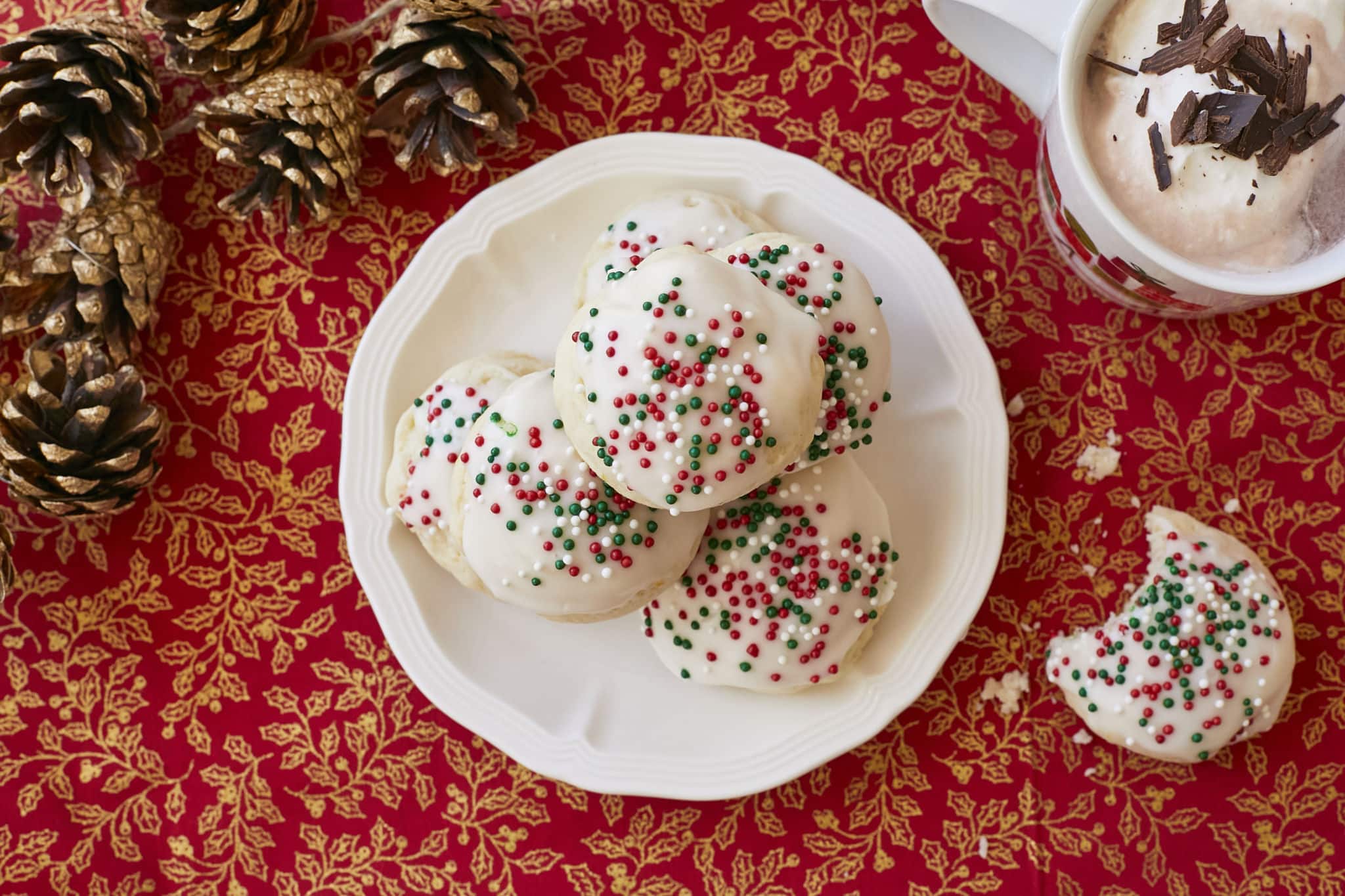A plate of homemade Italian Ricotta Cookies are presented on a red table cloth with gold accents next to a couple of pine cone. The Italian Ricotta Cookies are topped with a homemade vanilla glaze and red, green, and white Christmas sprinkles.